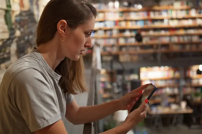How to Effectively Promote Your Book on Social Media Woman Using Smartphone in a Bookstore